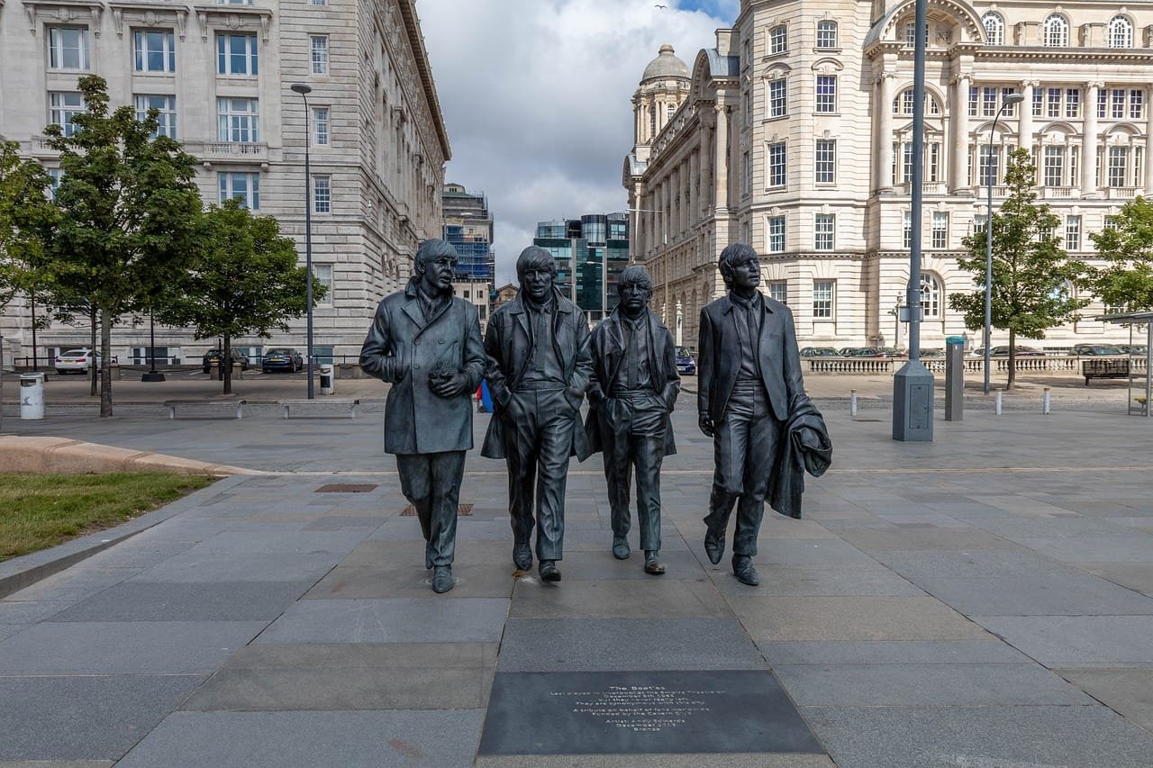 Bronze statue of The Beatles - John Lennon, Paul McCartney, George Harrison, and Ringo Starr - walking side by side at Pier Head in Liverpool, with historic buildings and a paved square in the background.